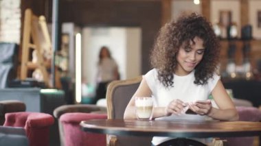 Smartphone'da mesajlaşma ve içtenlikle gülümseyen güzel İspanyol kadın. genç kız melez bir kafede kahve içer