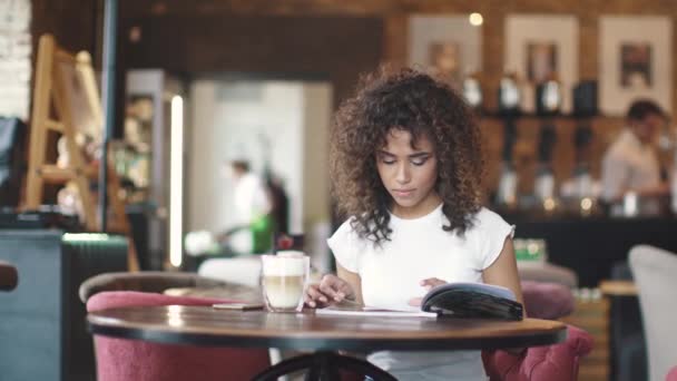 Chica joven mulato bebe café en un café y mira el menú. hermosa mujer hispana disfruta del café con leche en un acogedor café — Vídeo de stock