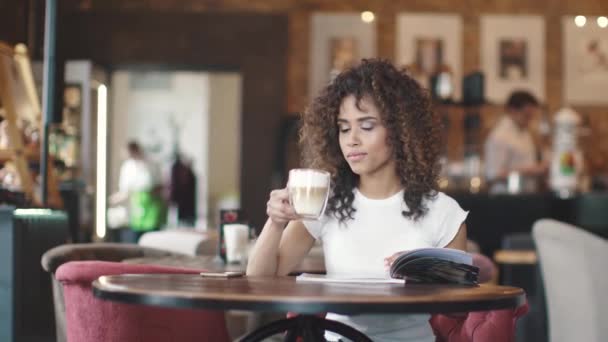 若い女の子のムラートは、コーヒーを飲みます。美しいヒスパニック系女性は、居心地の良いカフェでラテを楽しんでいます。多民族の外観を持つ少女の肖像画. — ストック動画