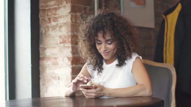 Atractiva chica hispana usando smartphone y sonríe en una cafetería . — Vídeos de Stock