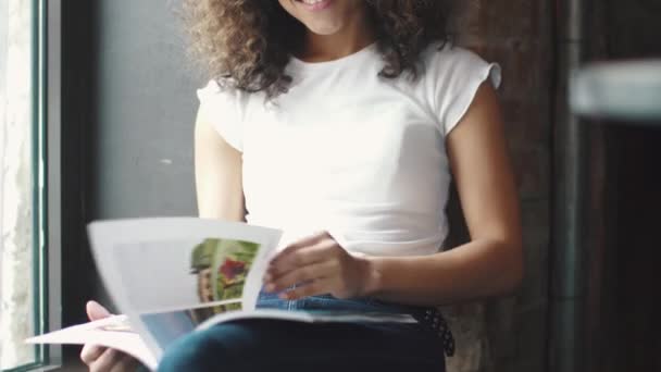 Девушка листает журнал сидя у окна в кафе — стоковое видео