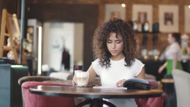 Flickan tittar på menyn i kaffehuset och dricker kaffe med mjölk. ung kvinna sitter vid ett bord på ett café — Stockvideo