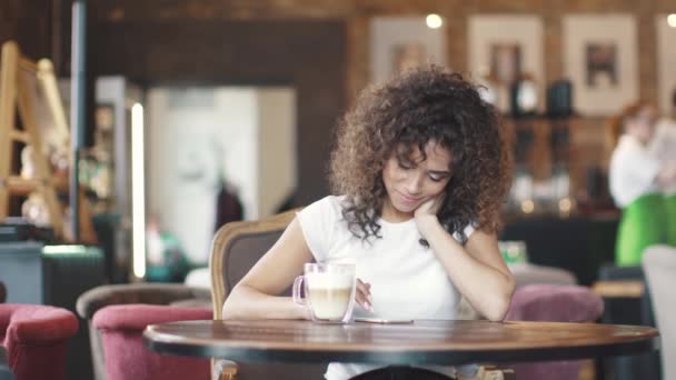 Ein attraktives hispanisches Mädchen beim SMS-Schreiben in einer mobilen Anwendung auf einem Smartphone in einem Café sitzend — Stockvideo
