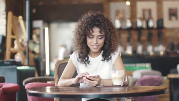 Retrato de uma bela menina hispânica em um café. mensagem de texto menina no smartphone e sorrisos — Vídeo de Stock