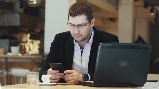 Mannen i kostym arbetar på en bärbar dator och mobiltelefon i ett café — Stockvideo