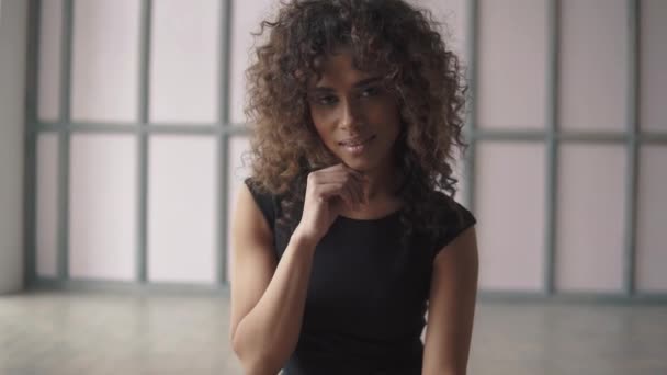 Retrato de cerca de una hermosa joven hispana con un vestido negro. chica de pelo rizado linda sonriendo y mirando a la cámara — Vídeos de Stock