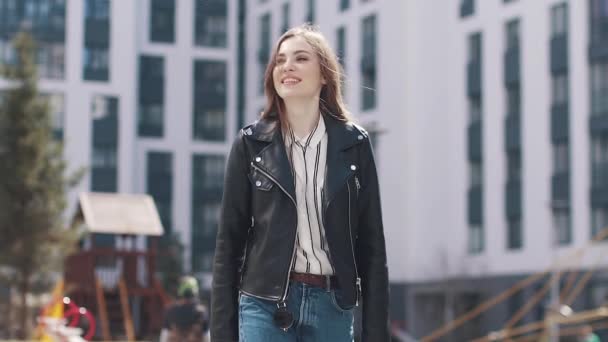 Ein junges modisches Mädchen spaziert in der Sonne durch die Frühlingsstadt. Porträt einer schönen jungen Frau vor dem Hintergrund moderner Architektur. Zeitlupe — Stockvideo