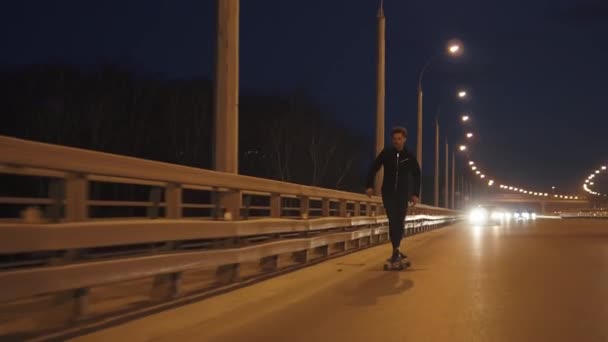 Парень ездит на длинном горке по городу ночью. Молодой человек катается на коньках по дороге вечером. замедленное движение — стоковое видео