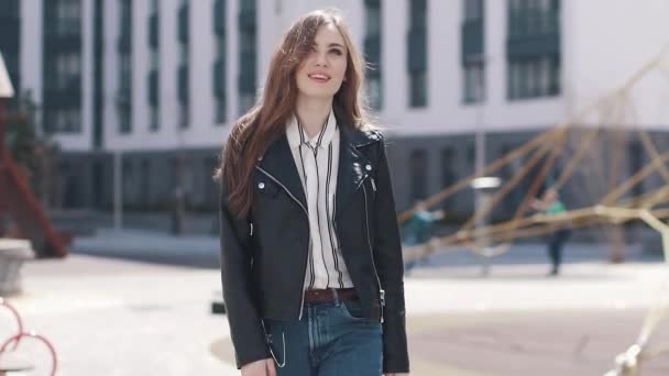 Uma jovem caminha pela cidade e, em movimento, tira o casaco. retrato de uma bela jovem mulher contra um fundo da arquitetura moderna. câmara lenta — Vídeo de Stock