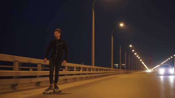 Ένας νεαρός άντρας που κάνει σκέιτ μπορντ σε μια πόλη το βράδυ στο οδόστρωμα. hipster τρόπο του ταξιδιού — Αρχείο Βίντεο