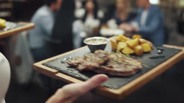 Camarero lleva un plato de carne a la mesa. la compañía de amigos reunidos para la cena en un café o restaurante — Vídeos de Stock