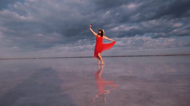 Ung kvinna tar en selfie samtidigt stående på stranden av en rosa sjö där himlen återspeglas — Stockvideo