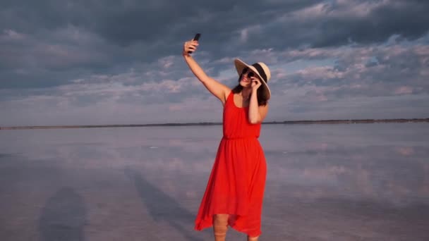 Jonge vrouw neemt een selfie in een prachtige ongebruikelijke plaats. meisje in een rode jurk, muts en zonnebril op een roze meer. — Stockvideo