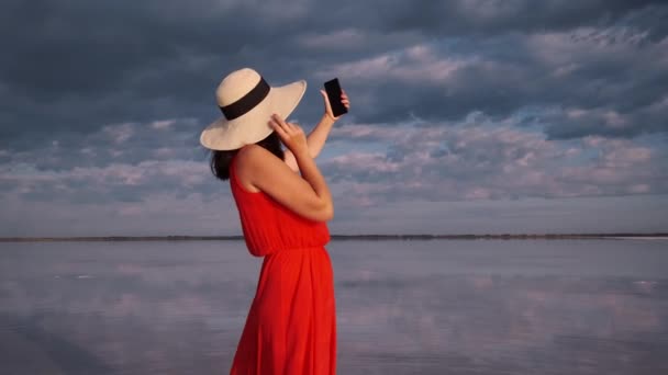 Young woman in a hat and sunglasses takes a selfie on a background of sky and clouds. Traveler takes photos on the phone camera — Stock Video