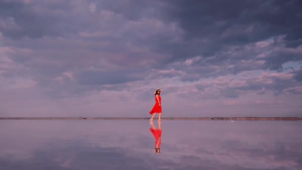 Jonge vrouw in een zwaaiende jurk loopt langs een roze meer waarin wolken worden gereflecteerd. — Stockvideo