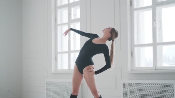 Mladá žena v černém obleku a kamaších se rozehřívá. dívka tanečnice dělá strečink v jasném studiu — Stock video