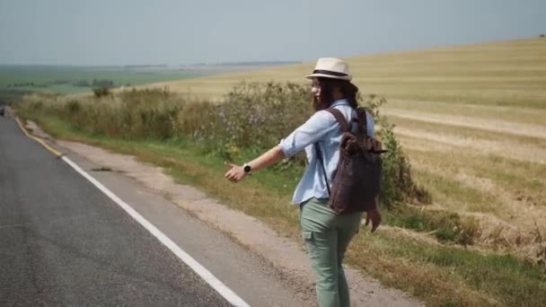 Resenär liftar vid vägkanten. turist med en ryggsäck och en hatt försöker stoppa bilen. — Stockvideo