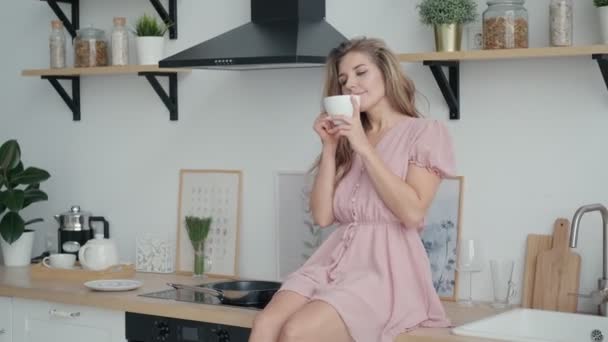Милая беззаботная девушка наслаждаясь утренним кофе и улыбаясь, сидя на столе на кухне — стоковое видео