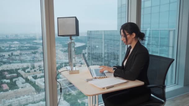 Zakenvrouw werkt in een modern kantoor gelegen op een hoge verdieping van een wolkenkrabber. aantrekkelijk meisje werken op een laptop terwijl zittend aan de tafel door een groot raam. — Stockvideo