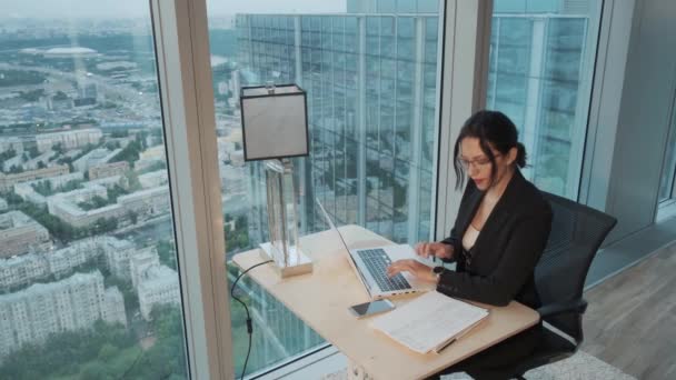 Νεαρή επιχείρηση γυναίκα που εργάζεται σε ένα φορητό υπολογιστή σε ένα μοντέρνο γραφείο που βρίσκεται στο ψηλό όροφο ενός ουρανοξύστη. — Αρχείο Βίντεο