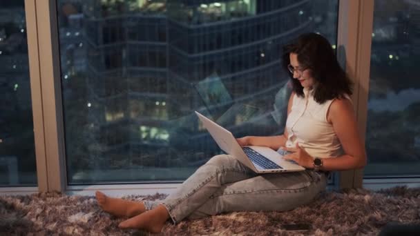 A young woman uses a laptop sitting comfortably by the window on a high floor — ストック動画