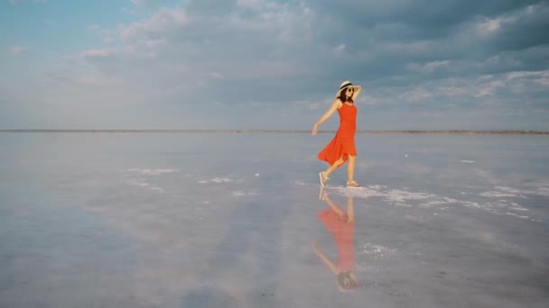 Tourist in a waving dress walks along a salt lake in which the sky is reflected — 비디오