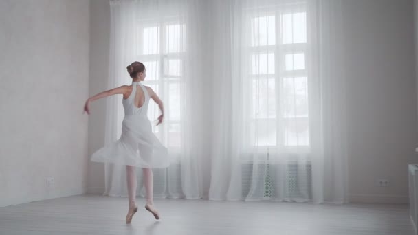 Balett dansare snurrar på tå i en snövit klänning i en ljus studio på bakgrunden av stora fönster — Stockvideo