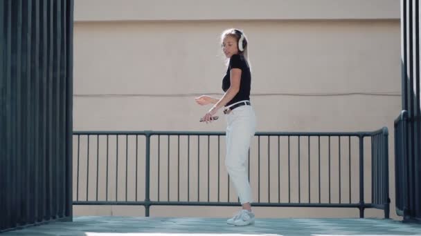 Молодая женщина весело танцует и прыгает высоко, слушая музыку в наушниках на открытом воздухе — стоковое видео