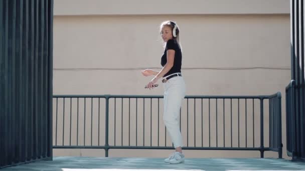 Young woman dances cheerfully and bounces high while listening to music on headphones outdoors — Stock Video