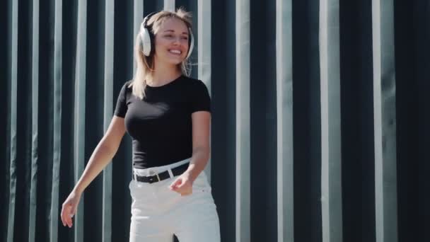 Menina sorridente ouve música em fones de ouvido e danças em um fundo de paredes pretas — Vídeo de Stock