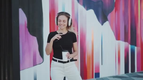 Vacker ung flicka rolig dans på bakgrunden av en färgad vägg. ung kvinna med ett charmigt leende lyssnar på musik på hörlurar — Stockvideo