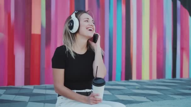 Menina despreocupada alegre ouvir música em grandes fones de ouvido ao ar livre contra uma parede multi-colorida — Vídeo de Stock