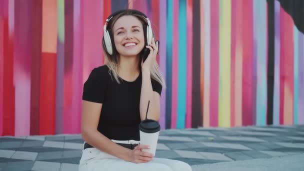 Retrato de uma menina atraente ouvir música com fones de ouvido e beber café ao ar livre — Vídeo de Stock