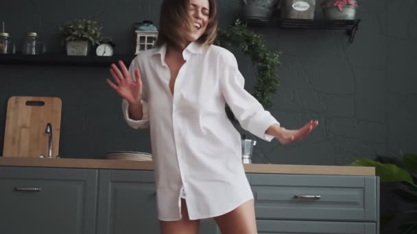 Young woman funny and carefree dancing in the kitchen in a white shirt. happy girl having fun and carefree dancing at home in the early morning — ストック動画