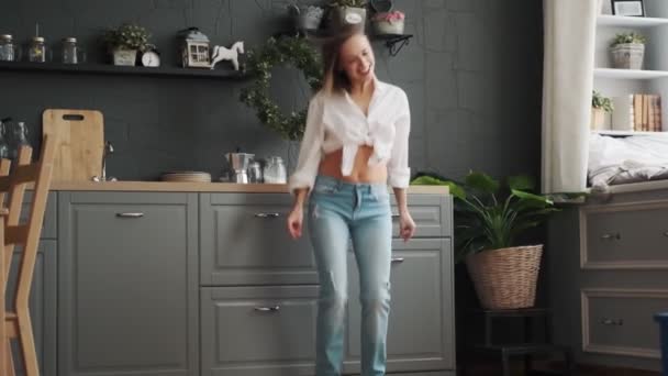 Молодая женщина веселится, танцуя в рубашке и джинсах дома. Девушка танцует смешно на кухне — стоковое видео