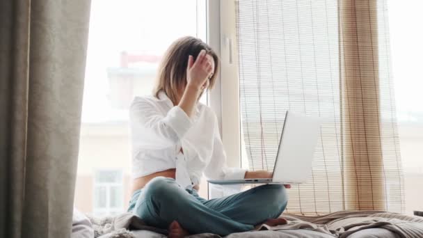 Молода жінка працює на ноутбуці, сидячи зручно на підвіконні вдома. дівчина з ноутбуком посміхається і смс — стокове відео