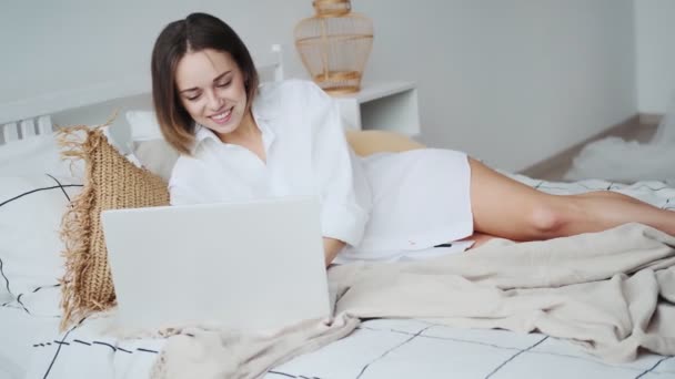 Young woman uses a laptop while lying in bed. girl working on a laptop in the morning, comfortably sitting in bed — Stock Video