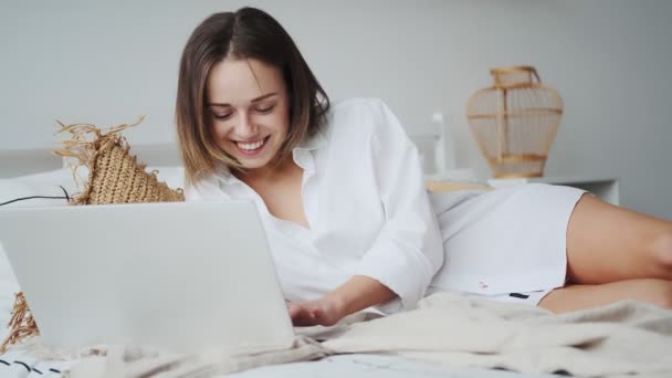 Lycklig kvinna med laptop liggande i sängen och leende. flicka i en mans skjorta arbetar på en bärbar dator tidigt på morgonen — Stockvideo