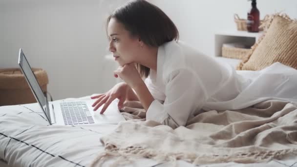 Leuke vrouw gebruikt een laptop terwijl ze in bed ligt en lacht. meisje in een mans shirt werken op een laptop in de vroege ochtend — Stockvideo