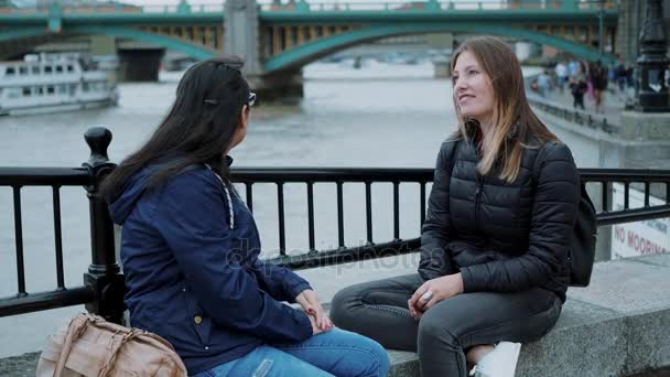 Two girls sit at the banks of River Thames and have small talk — Stock Video