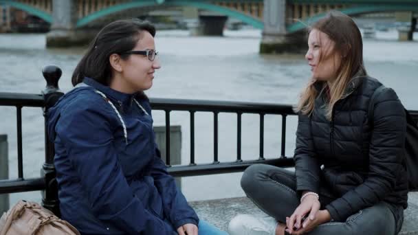 Two girls sit at the banks of River Thames and have small talk — Stock Video