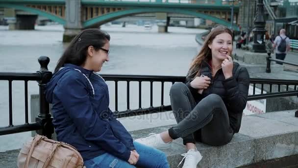 Dos chicas se sientan a orillas del río Támesis y tienen una pequeña charla — Vídeo de stock