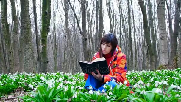 Une belle fille intelligente lit un livre intéressant dans une forêt printanière pleine de gouttes de neige en fleurs — Video