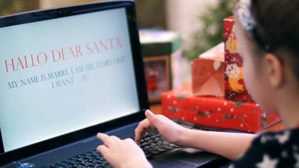 Niedliche blonde Mädchen in einem schönen Kleid druckt einen Brief an den Weihnachtsmann auf einem Laptop, in der Nähe eines Weihnachtsbaums, dekoriert mit schönen Weihnachtslichtern. Spielzeug und Bälle — Stockvideo