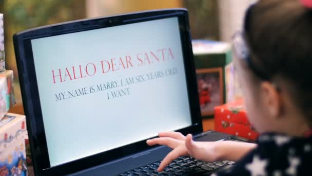 Jolie fille blonde dans une belle robe imprime une lettre au Père Noël sur un ordinateur portable, près d'un arbre de Noël, décoré de belles lumières de Noël. jouets et balles — Video