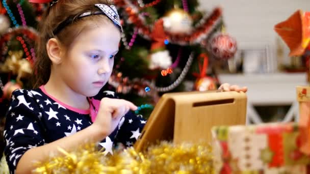 Linda chica rubia en un hermoso vestido es la impresión de una carta a Santa en una tableta, cerca de los regalos y un árbol de Navidad, decorado con hermosas luces de Navidad. juguetes y pelotas — Vídeo de stock