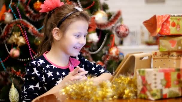 Linda chica rubia, con un lazo rosa en el pelo, en un hermoso vestido está viendo algo en una tableta, cerca de los regalos y un árbol de Navidad, decorado con hermosas luces de Navidad. juguetes y pelotas — Vídeo de stock