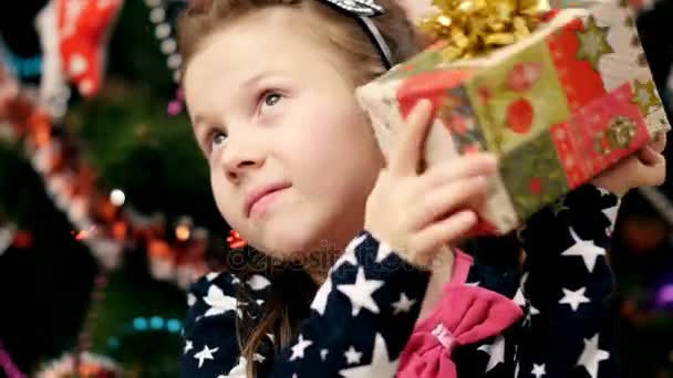Güzel sarışın kız güzel şık elbiseli, saçında pembe bir yay ile merakla onun Noel hediyesine arar, sonra onun kulağına sallıyor — Stok video