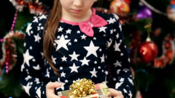 Bella ragazza bionda con un fiocco rosa tra i capelli, in un bel vestito elegante, guarda con curiosità il suo regalo di Natale, poi lo scuote vicino al suo orecchio — Video Stock