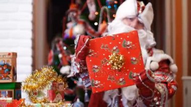 Canta Klaus, Noel Baba, baba Frost altın bir yay ile bir kırmızı kağıt ambalajı içinde büyük bir Noel hediyesi mittens elinde tutar, eliyle ok gösterir, arka planda bir Noel ağacı.